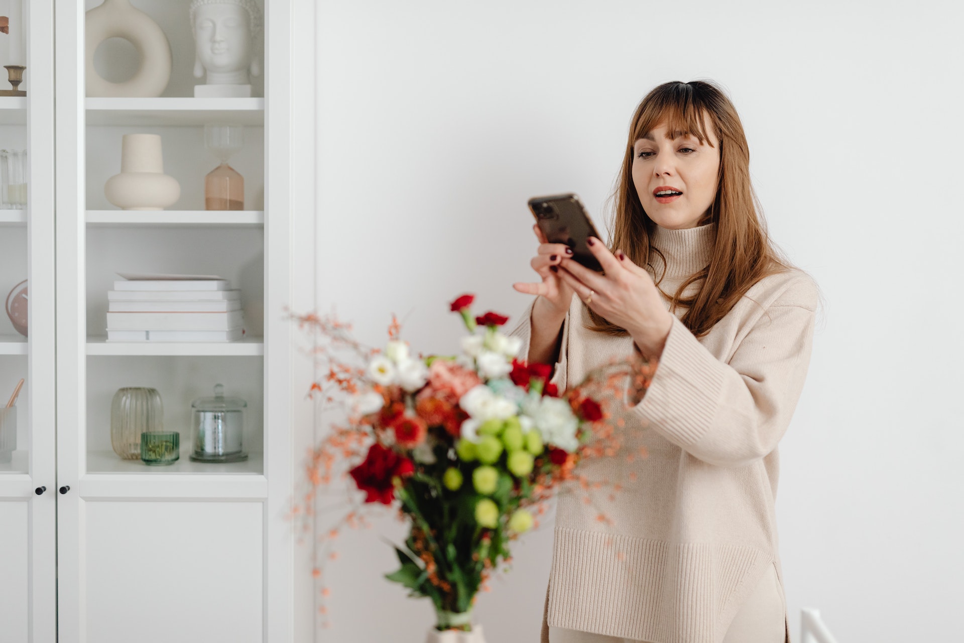 A photo of a woman taking a photo of a bunch of flowers in a vase on a table.