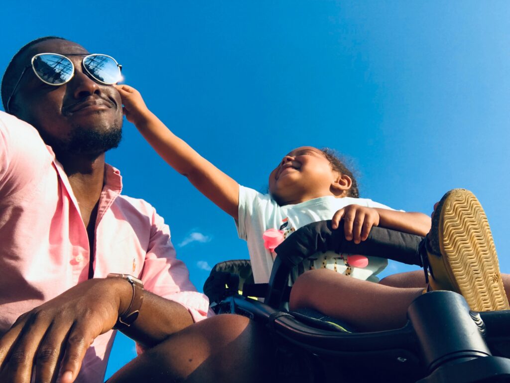 Father playing with his toddler daughter on vacation, laughing as she tries to remove his sunglasses from his face